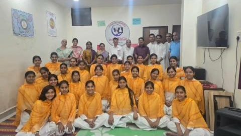 Group photograph — about thirty students in orange seated and standing before the Sarvarthasiddhi emblem, with the scholars and guests behind them