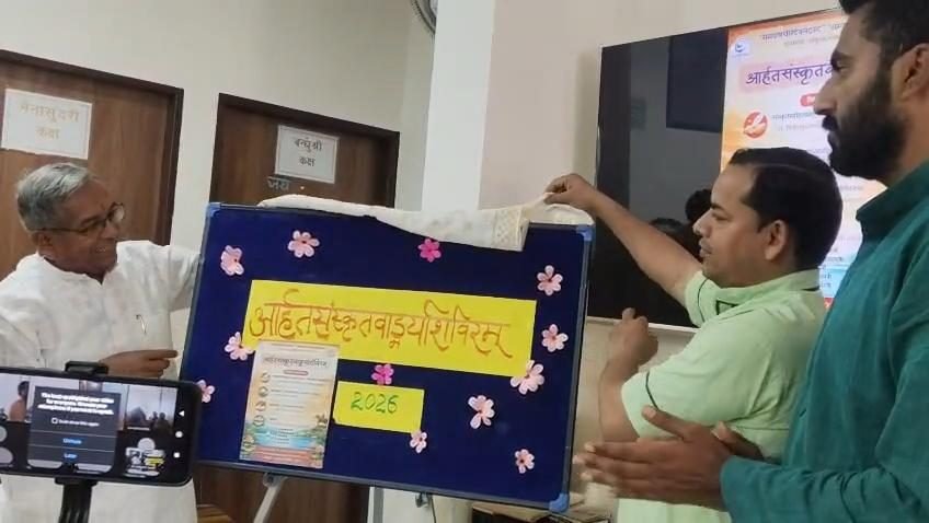 Two men drawing the cloth from a board reading आर्हतसंस्कृतवाङ्मयशिविरम् 2026 while an elder looks on