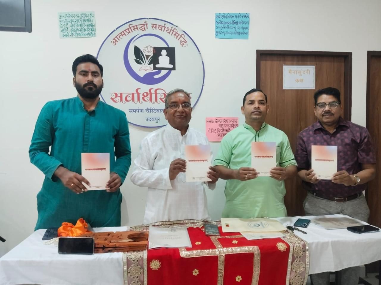 Four men holding copies of a newly released Sanskrit booklet before the Sarvarthasiddhi emblem