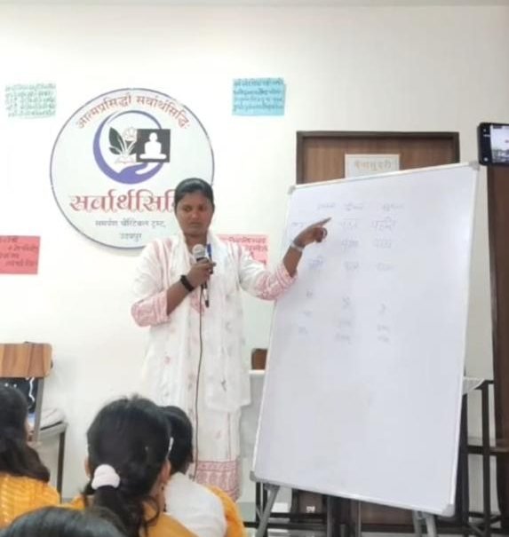 A teacher at a whiteboard, microphone in hand, pointing to a table of Sanskrit word forms while the students watch