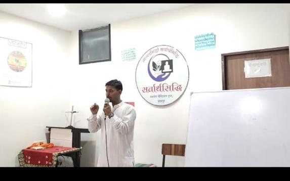 A young man with a microphone speaking in front of the Sarvarthasiddhi emblem and a whiteboard