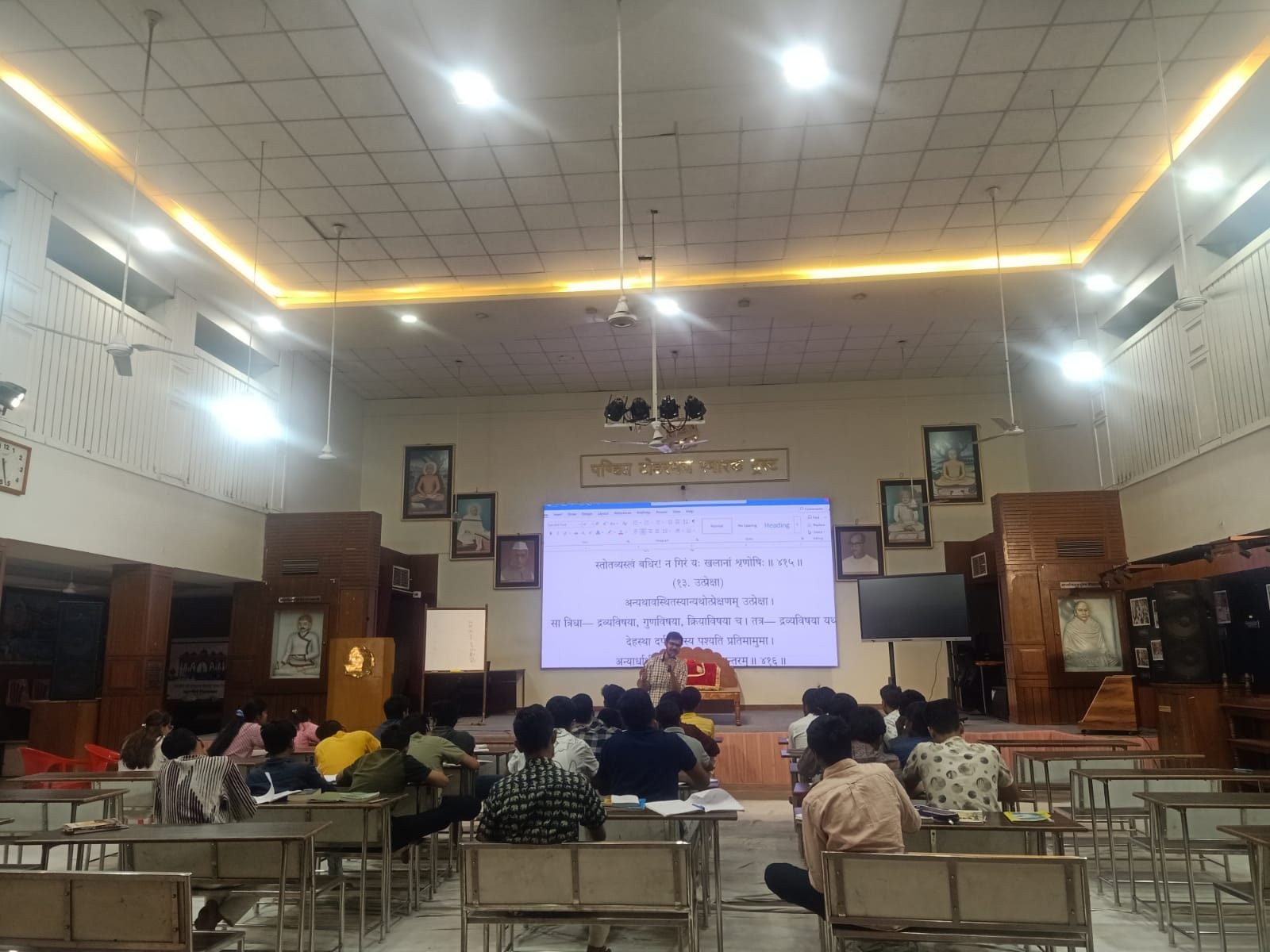 The workshop hall with participants seated at desks and a speaker at the front, a Sanskrit text projected on the screen