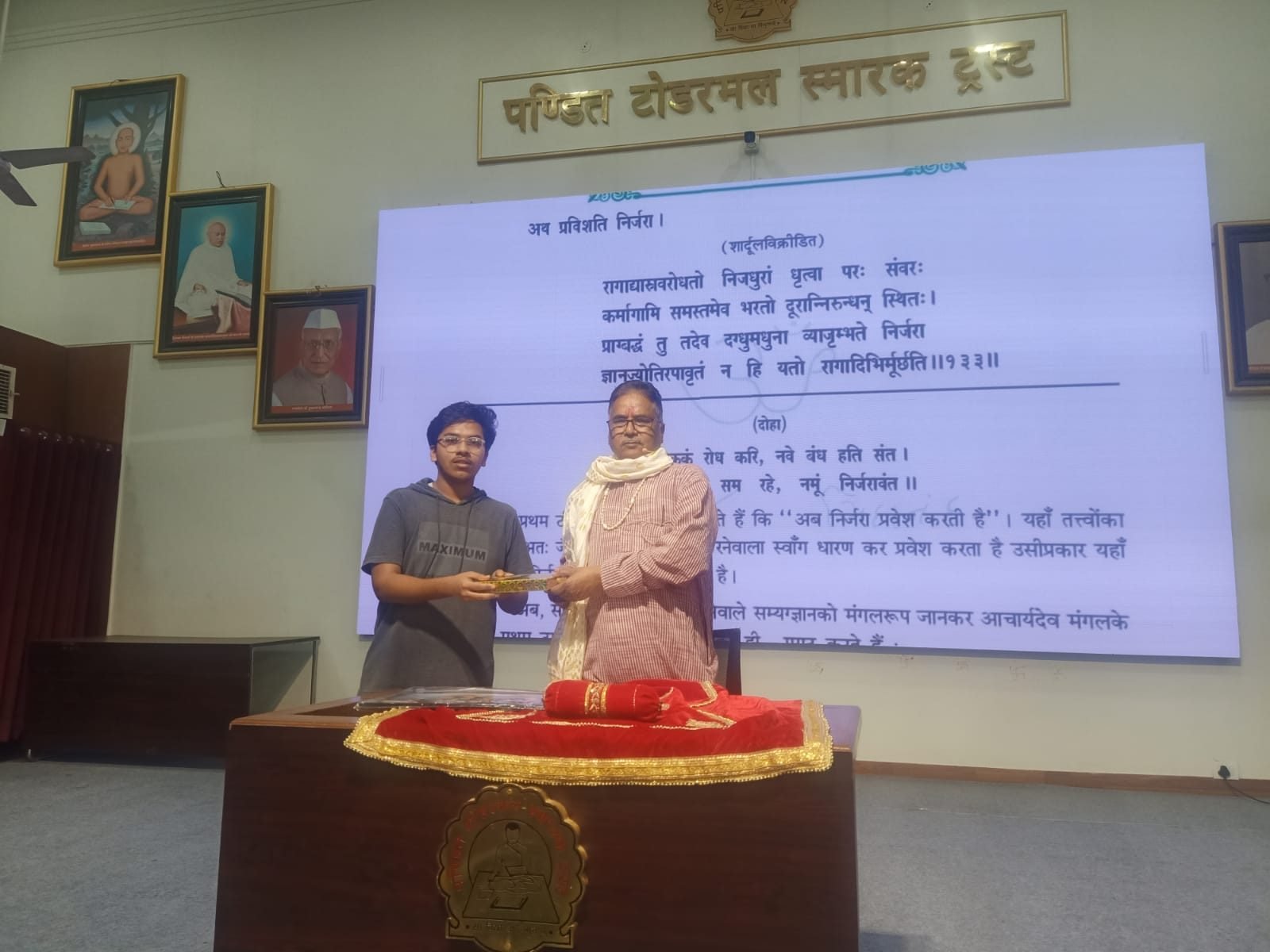 A participant offering a token to a senior scholar at the podium, with a Sanskrit verse projected behind them