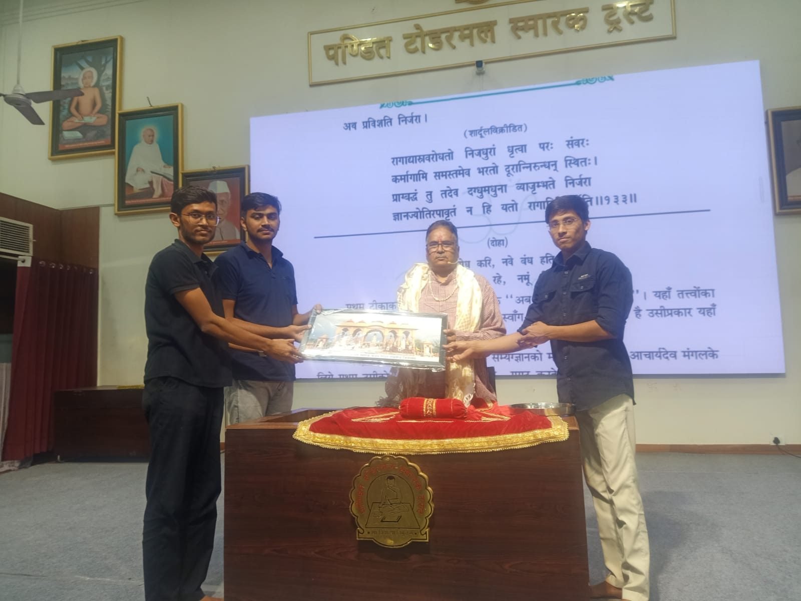Two participants presenting a framed memento to a guest, the projected Sanskrit verse behind them