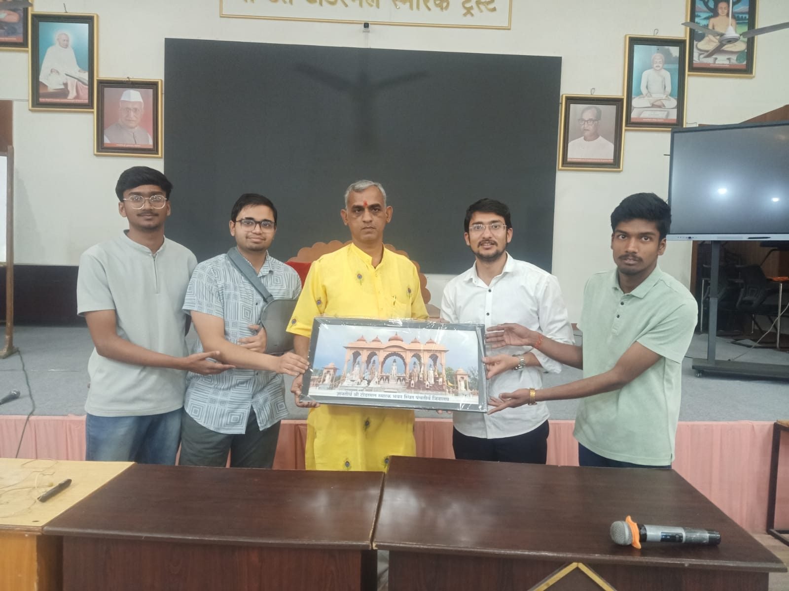Four participants standing with a guest, holding a framed memento between them