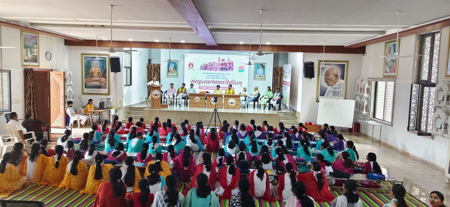 A wide view of the hall during the inaugural session, the participants seated on the floor before the dais and the camp banner projected behind it