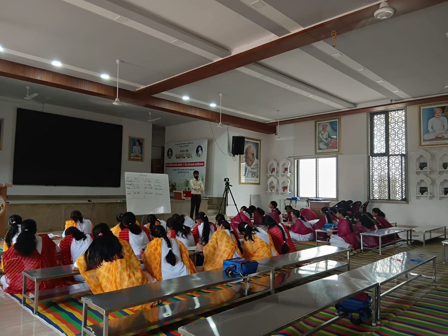 Participants seated at low desks facing a teacher who stands beside a whiteboard carrying a hand-drawn table