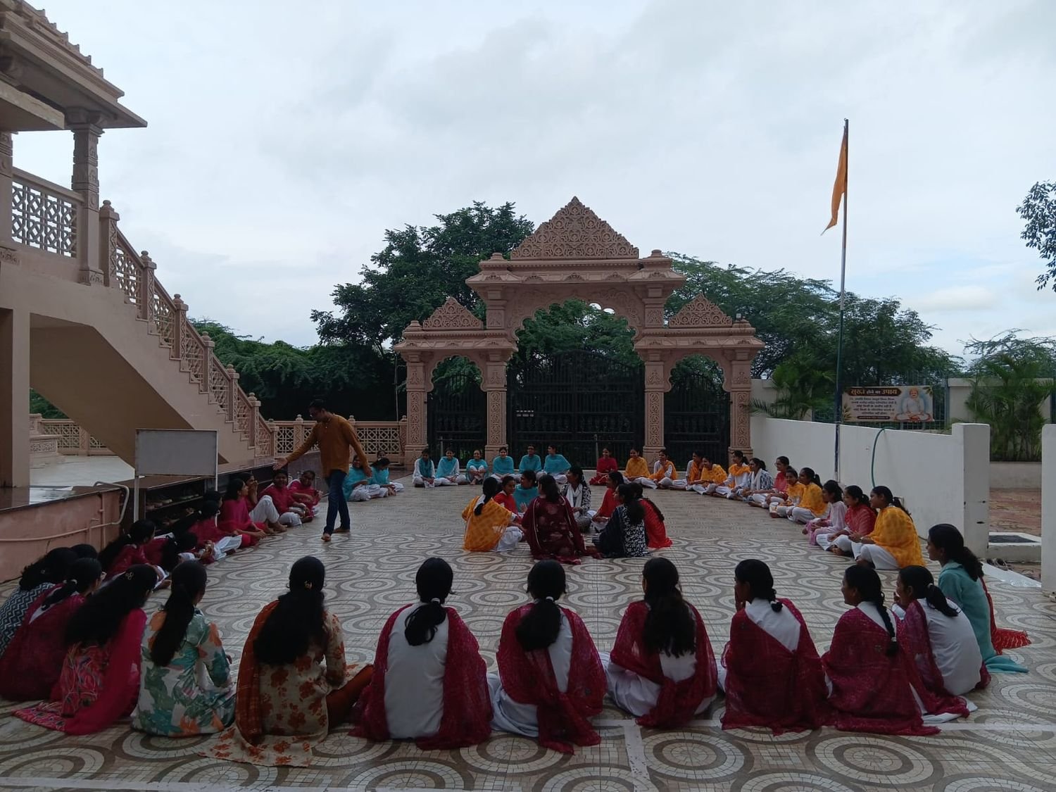 Participants seated in a large circle on a paved terrace before the carved stone gateway of Shashwat Dham, a teacher standing among them