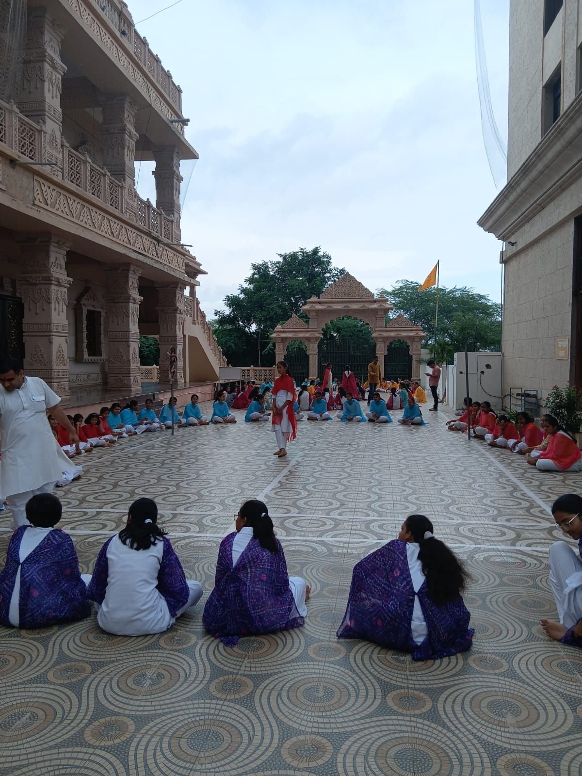 Participants seated in a wide circle on the patterned courtyard floor between the carved buildings of Shashwat Dham