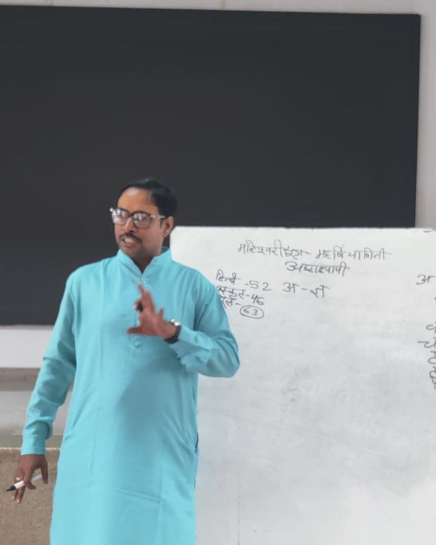 A teacher in a blue kurta standing beside a whiteboard on which the Maheshvara sutras and the letters of the alphabet are written