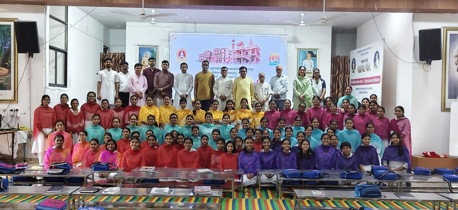 The participants of the Sanskrit Sambhashan Shivir seated in rows in the hall at Sanskar Tirth, Shashwat Dham, with the scholars and guests of the camp standing behind them before the projected announcement